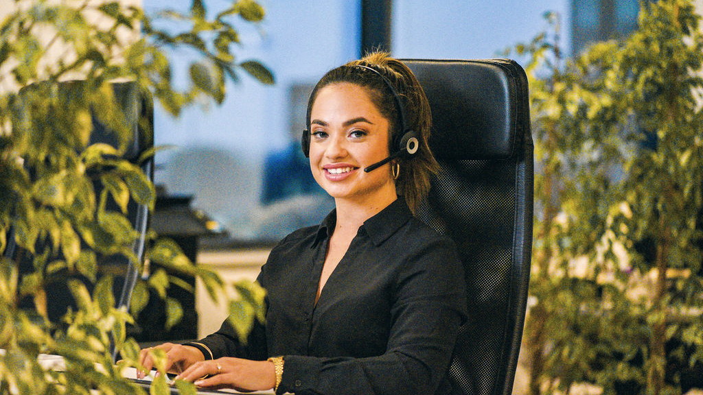 Recruiter met headset achter haar computer op kantoor, tussen groene planten, in gesprek met een kandidaat.